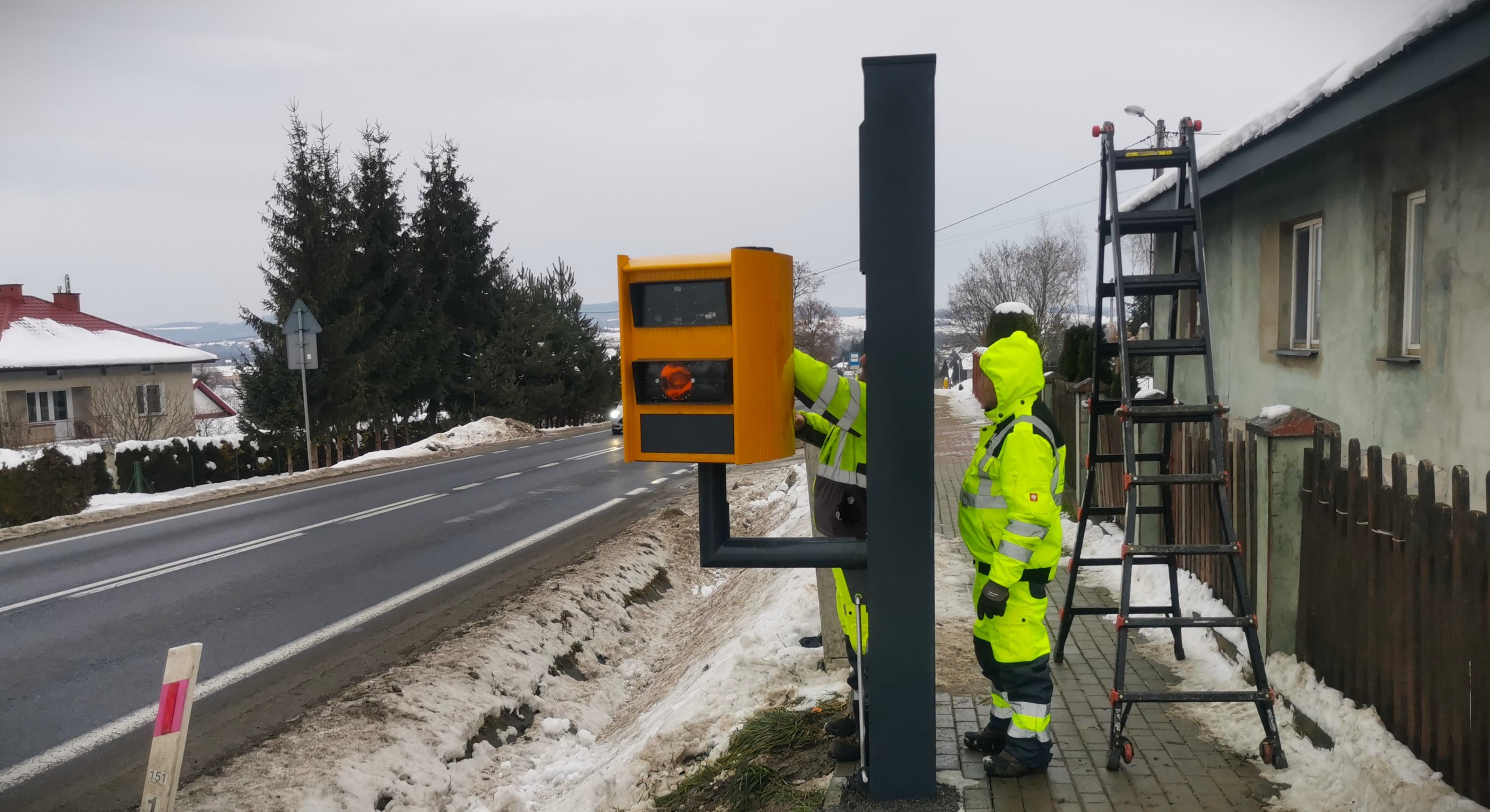 Dwóch mężczyzn w odblaskowych strojach stoi przy maszcie fotoradarowym